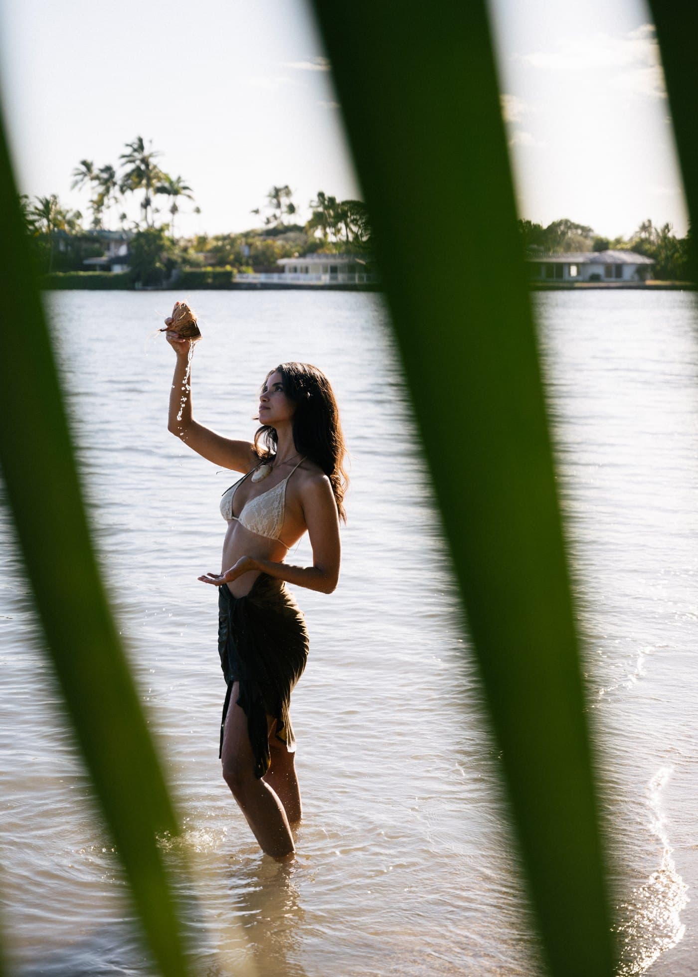 Model in the water at golden hour holding a coconut, framed by palm fronds
