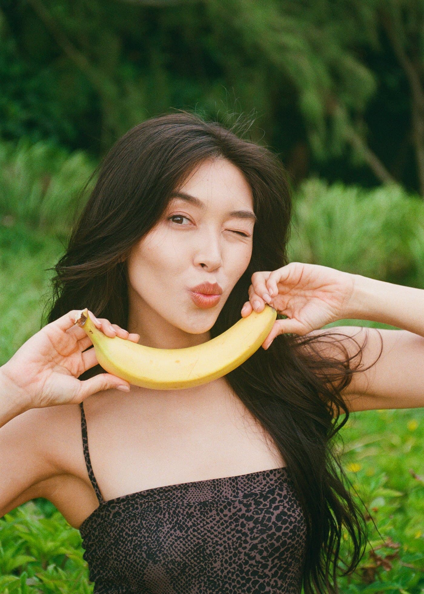 Model winking, holding a banana like a smile, green foliage behind